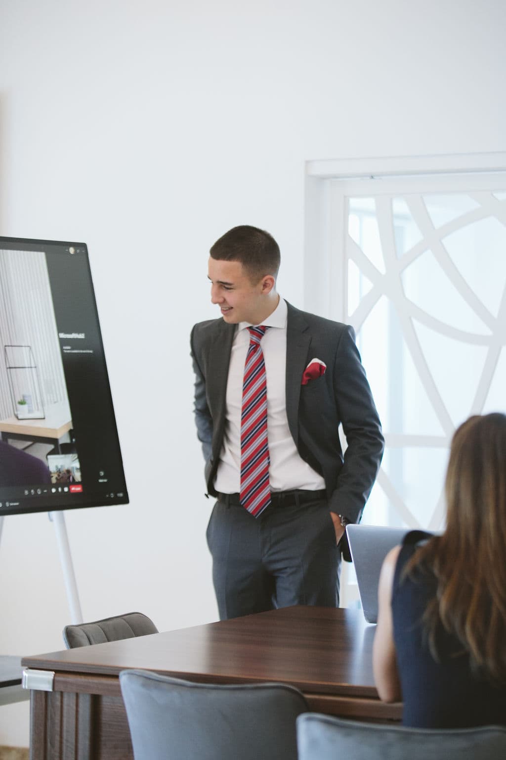 Smiling man in a suit and striped tie stands during a presentation in a modern office.