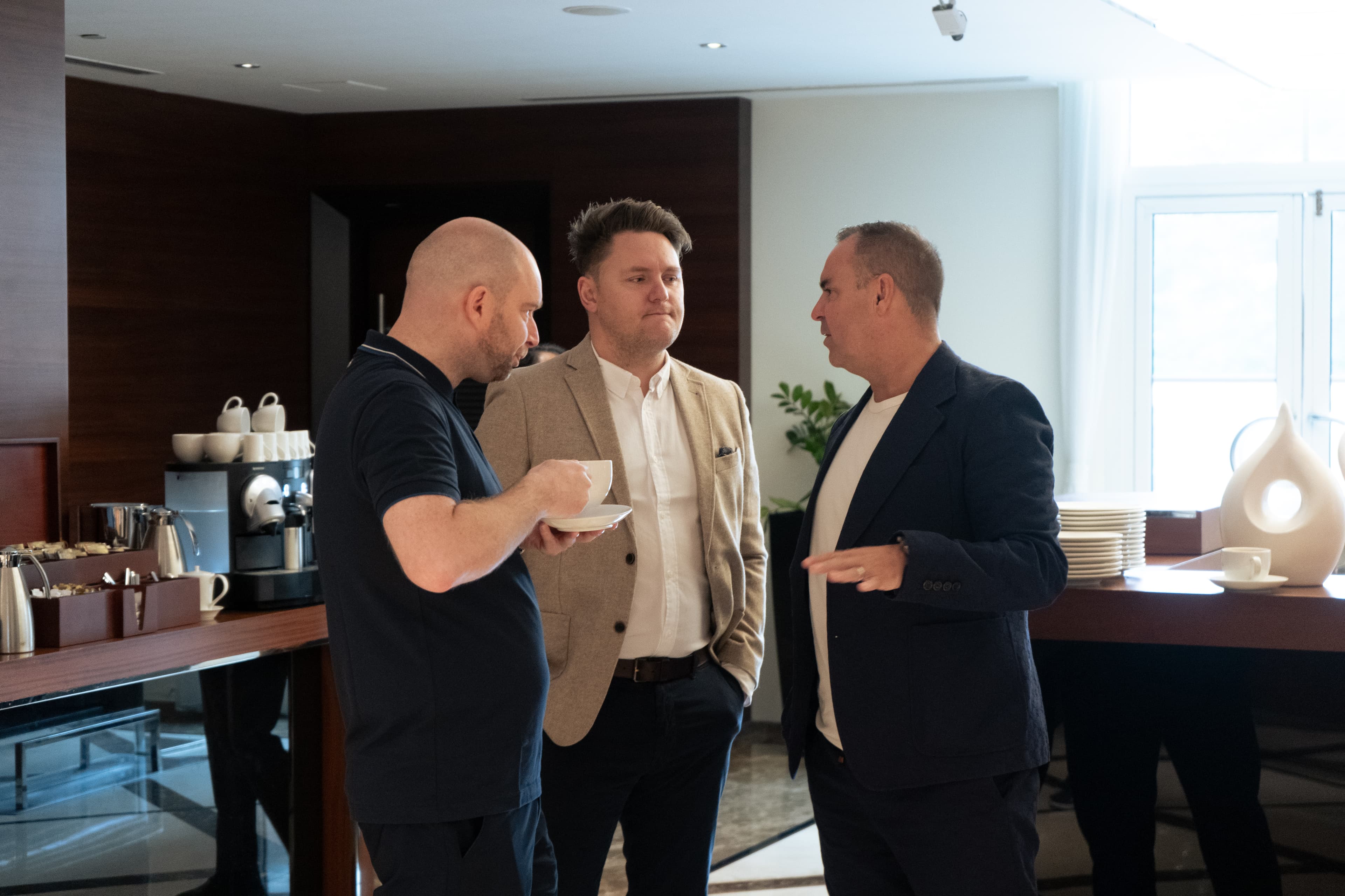 Three men in business casual attire talk and hold coffee in a modern, professional lounge.