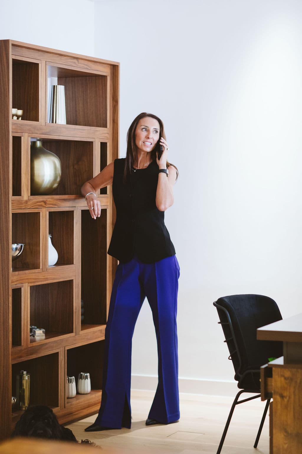 Woman in black vest and blue trousers talking on phone beside a wooden bookshelf.