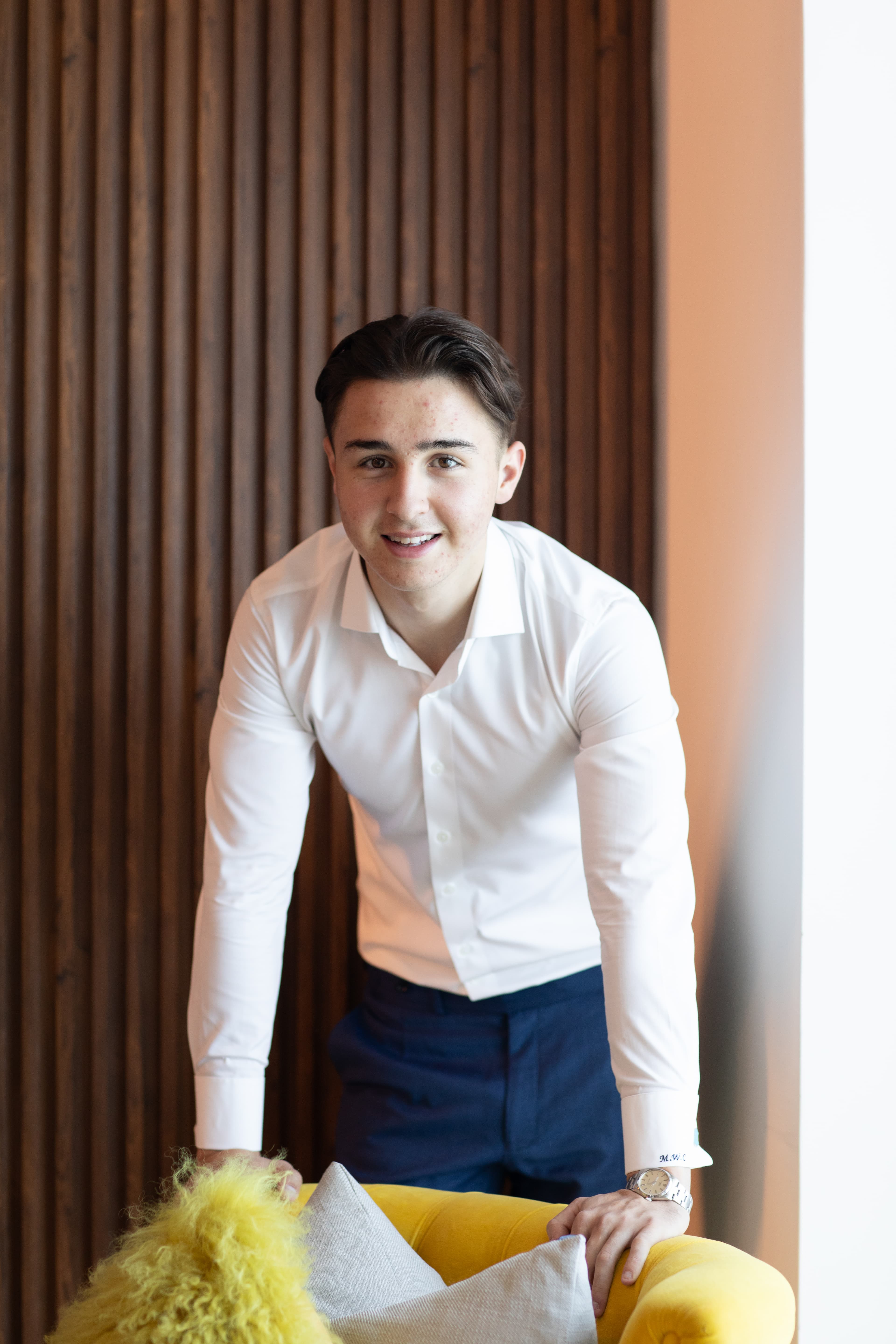 Smiling young man in white shirt leaning over a yellow armchair against slatted wood.