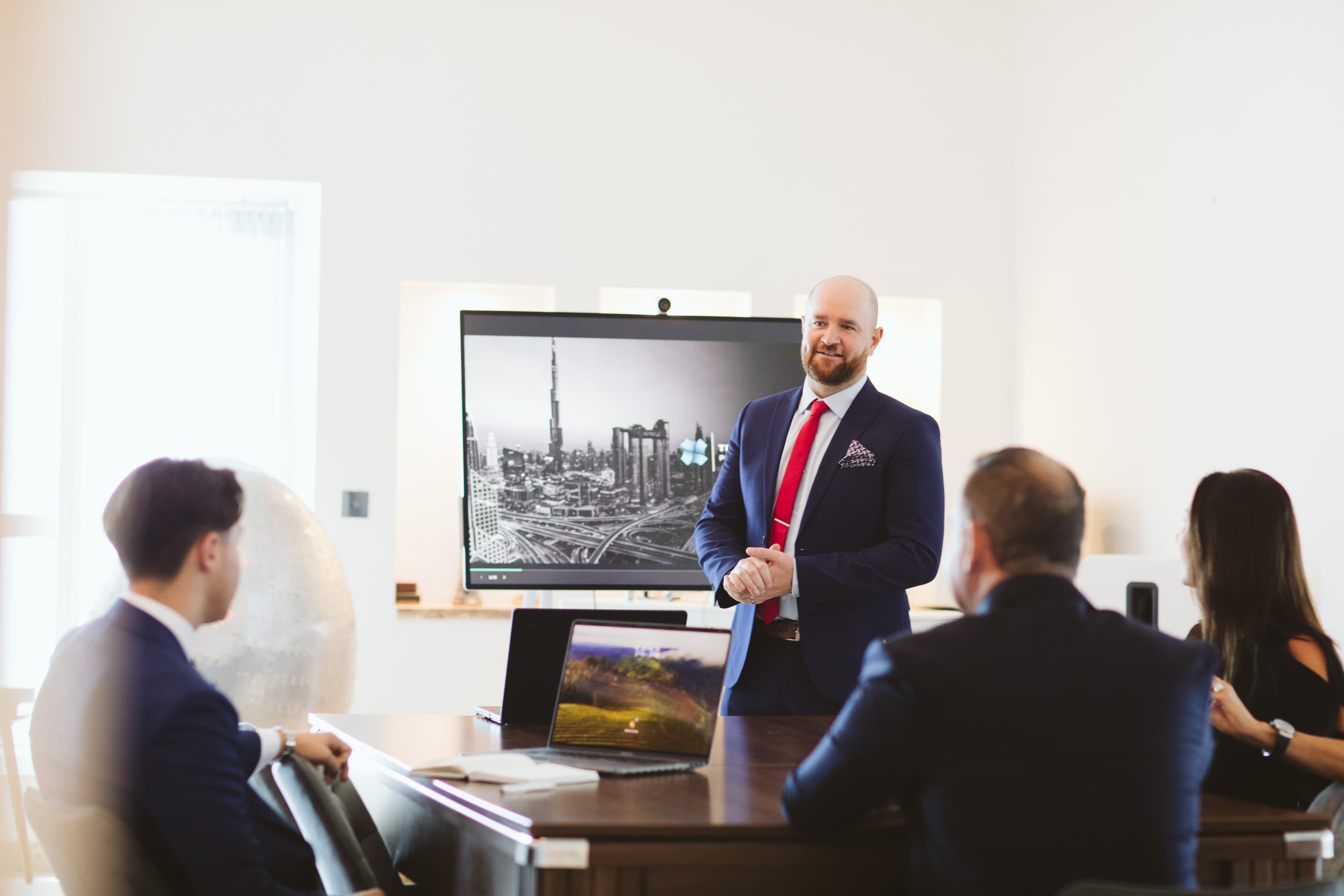 Man in a suit and red tie presenting to colleagues in a bright, modern office.