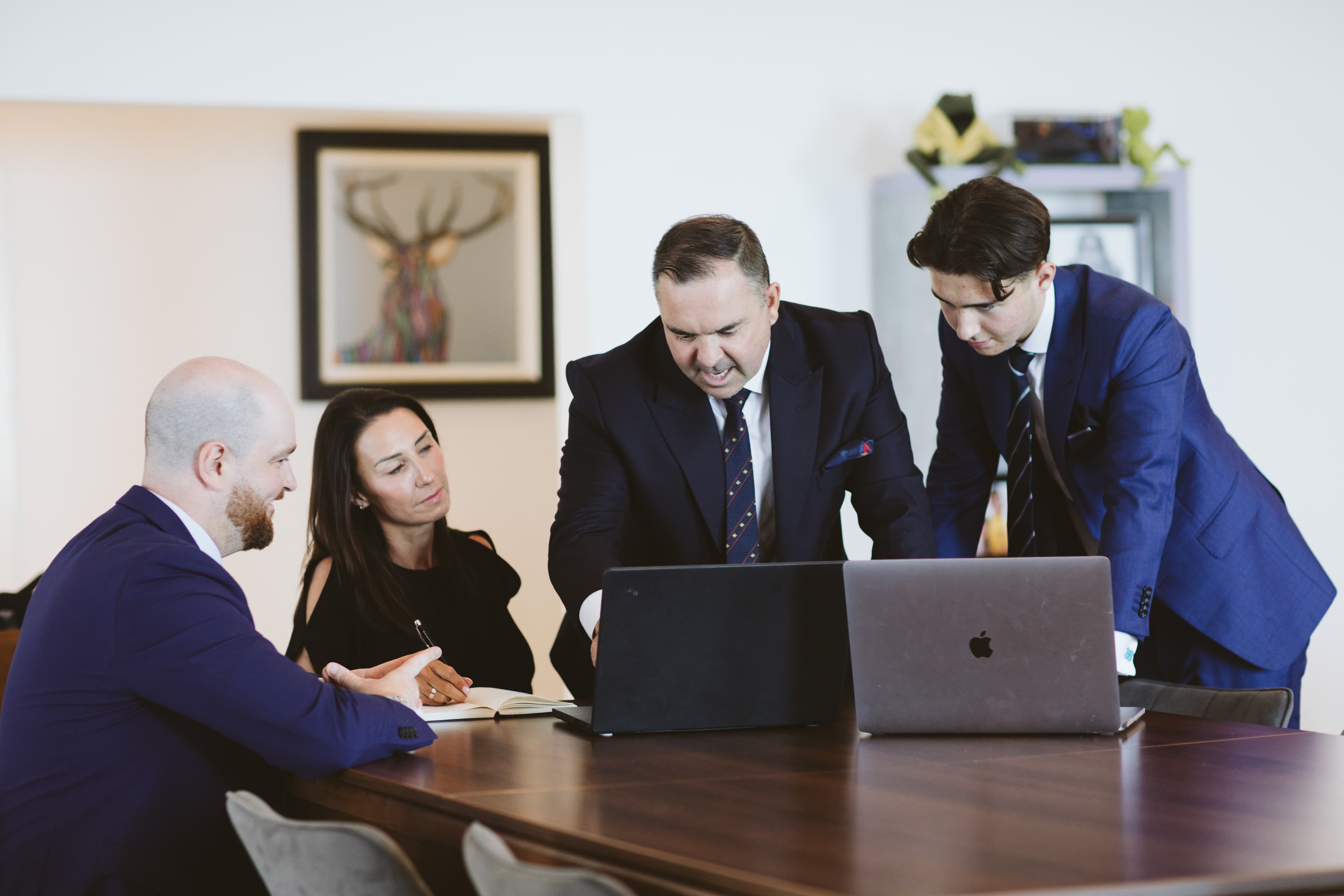 Four business professionals in suits discuss work while looking at laptops around a wooden table.